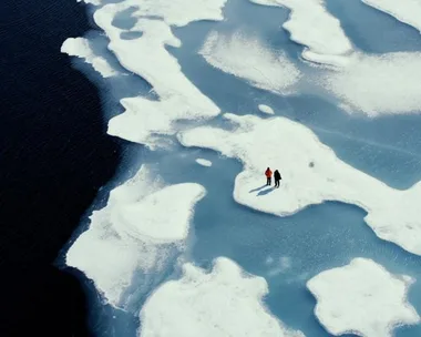 Aerial view of two people standing on a large ice formation surrounded by dark water.