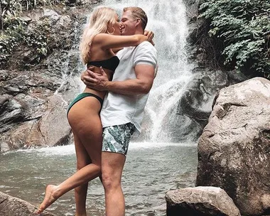 A couple embraces and kisses near a waterfall, with lush greenery surrounding them.