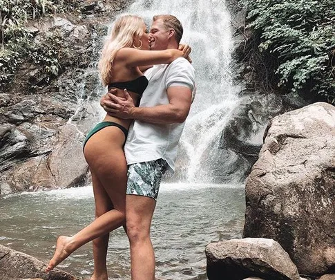 A couple embraces and kisses near a waterfall, with lush greenery surrounding them.