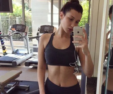 The image shows a woman in workout attire taking a selfie in a gym, with treadmills and exercise equipment in the background.