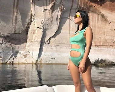 Kourtney Kardashian in a green cutout swimsuit stands on a boat with rocky cliffs in the background.