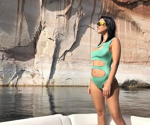 Kourtney Kardashian in a green cutout swimsuit stands on a boat with rocky cliffs in the background.