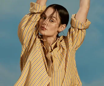 A woman in a striped yellow shirt poses with arms raised against a sunny sky backdrop.