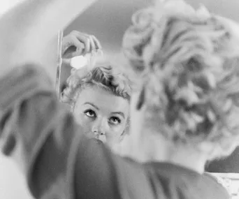 A woman styling her blonde curls in front of a mirror, captured in a vintage black-and-white photograph.