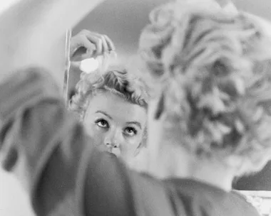 A woman styling her blonde curls in front of a mirror, captured in a vintage black-and-white photograph.