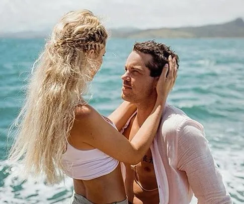 A couple embraces by the sea, with the woman holding the man's face and the ocean in the background.