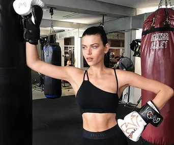 Woman in sports bra and boxing gloves standing in gym, resting hand on punch bag.