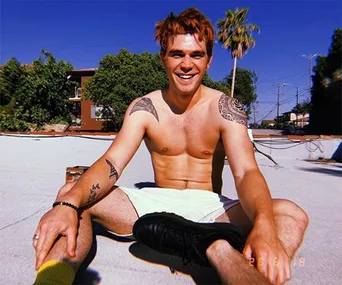 A shirtless man with tattoos sits outdoors on a sunny day, smiling and touching his sneakers.