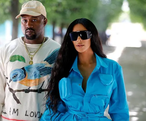 Kim Kardashian in blue jumpsuit and sunglasses, standing with Kanye West in a shirt with a bird graphic in a tree-lined path.