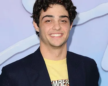 A young man with dark, curly hair smiling in front of a blue and white background, wearing a black blazer over a yellow T-shirt.