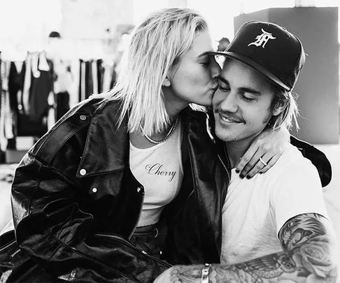 Black and white photo of a woman kissing a smiling man on the cheek. They're both seated; he wears a cap and she a shirt reading "Cherry."