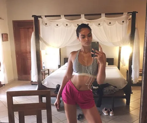 Woman in athletic attire takes a mirror selfie in a hotel room, wearing a sports bra, shorts, and earphones.