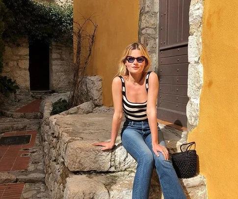 Woman in striped top and jeans sits on stone ledge, wearing sunglasses, with yellow wall background.