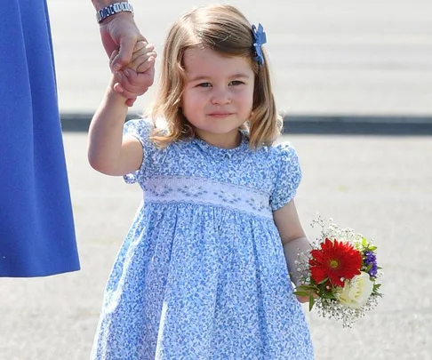 A young girl wearing a blue floral dress with a bow in her hair holds a small bouquet and an adult's hand.