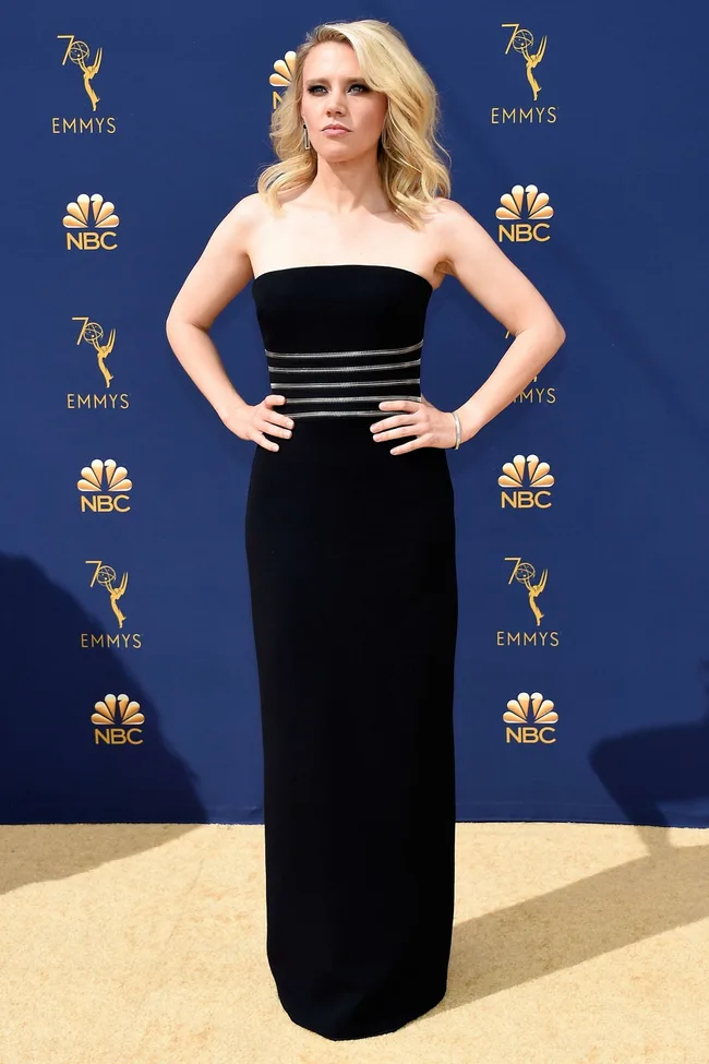 Person in a black strapless gown poses confidently on a light-colored carpet at an Emmy Awards event.