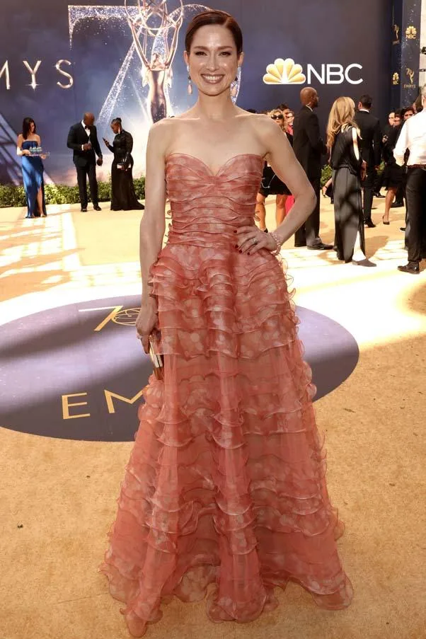 Person in a ruffled, strapless coral gown on the red carpet at the Emmy Awards, standing with hands clasped.