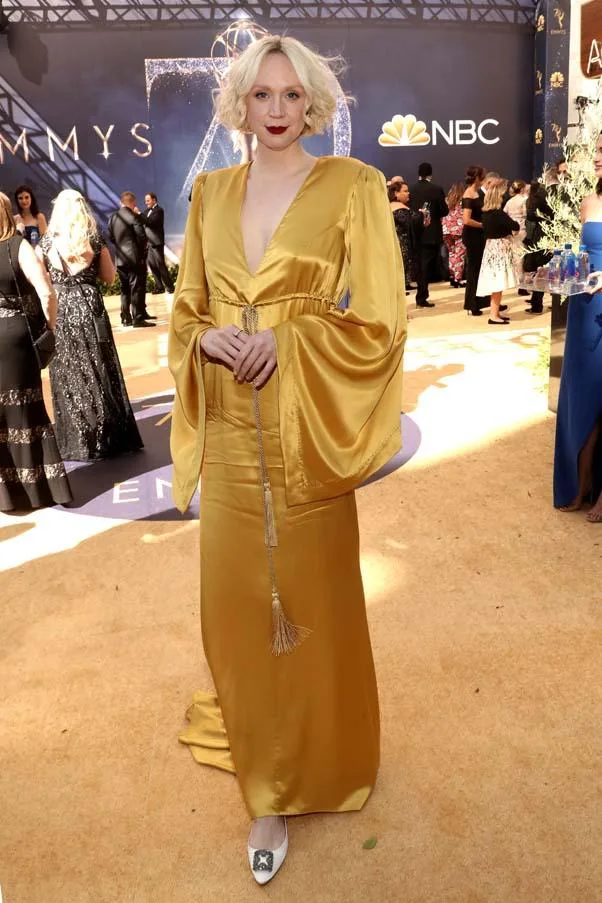 Person in a flowing yellow gown attends an award event, standing on a gold carpet with "Emmys" backdrop.