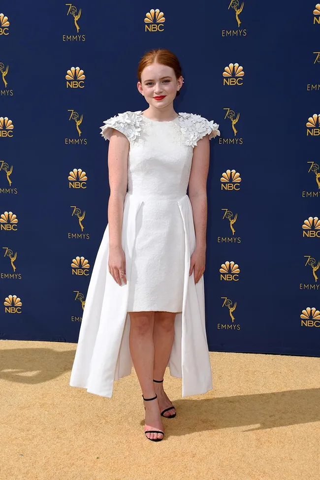 A woman in a white dress with floral shoulders at the 70th Emmy Awards on a beige carpet against a blue NBC backdrop.