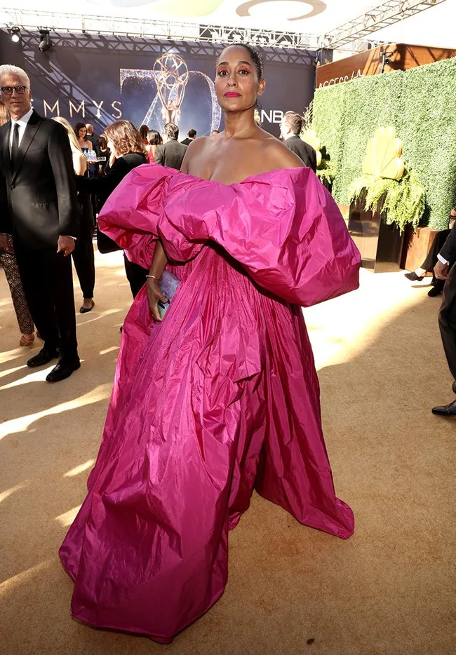 Woman in a vibrant pink gown on a red carpet, surrounded by people and Emmy-themed decor in the background.