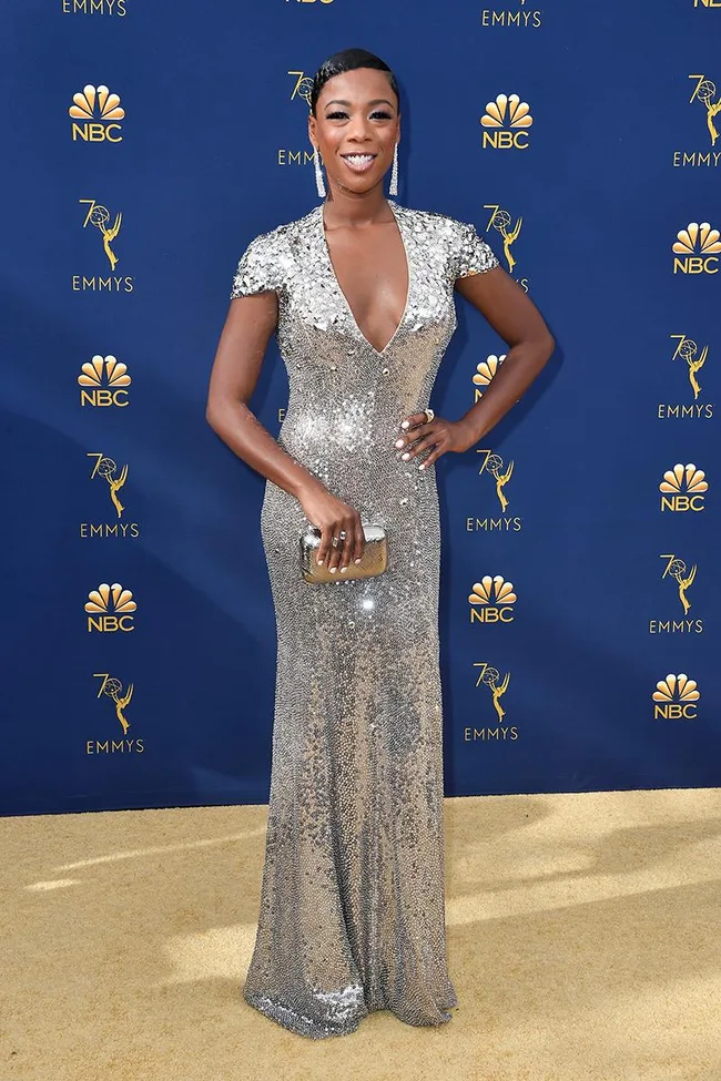 Person in a sparkling silver gown holding a clutch, on the red carpet at the 70th Emmy Awards, NBC backdrop behind.