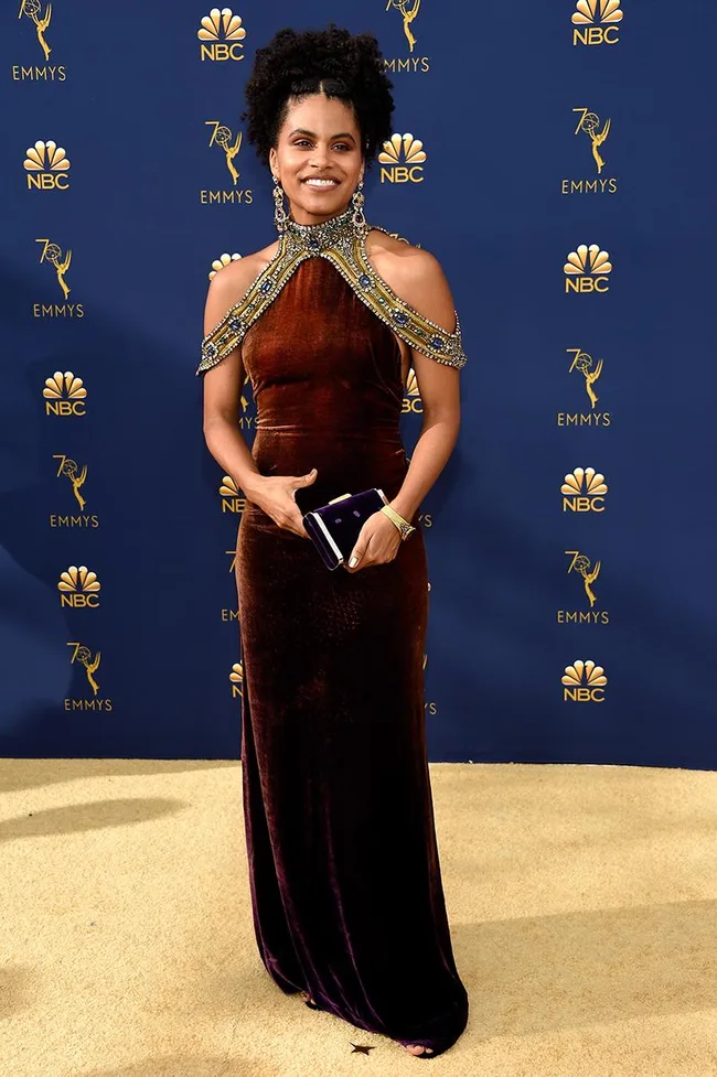 Actor in brown beaded gown at 70th Emmy Awards, holding a clutch, in front of NBC and Emmy logos on a blue background.