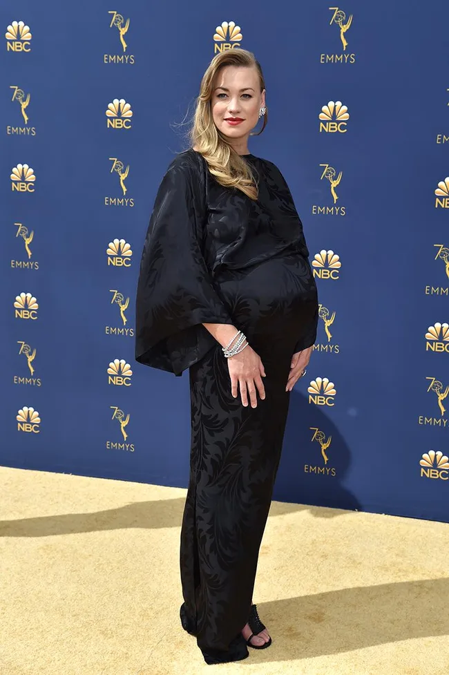 Woman in a long black dress on the Emmys red carpet, posing sideways with a hand on her belly.