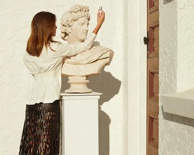 A woman holds Dior Joy perfume in front of a classical bust statue outside, against a textured white wall.