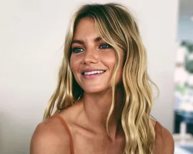 Blonde woman with wavy hair smiling, wearing a thin-strapped top, against a light blurred background.