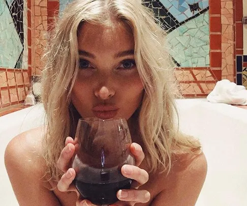 Woman with long blonde hair in a bathtub holding a glass of red wine, with colorful tiles in the background.