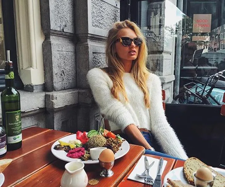 Woman in sunglasses and cozy sweater sitting at an outdoor table with a variety of food dishes.