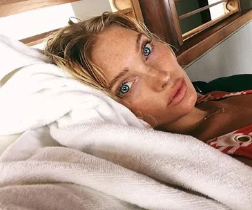 A woman with light hair and blue eyes lying down on white sheets, looking into the camera with a neutral expression.