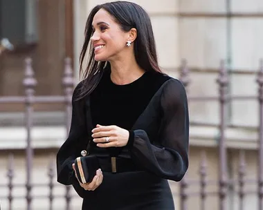 Woman in black dress with sheer sleeves, holding a clutch, smiles while walking near a building with stone architecture and metal fencing.