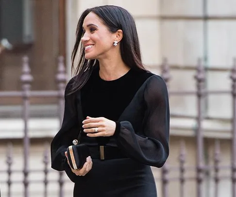 Woman in black dress with sheer sleeves, holding a clutch, smiles while walking near a building with stone architecture and metal fencing.