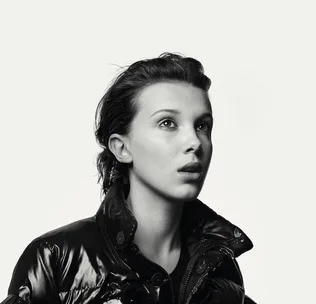 Black and white image of a young person with short hair, wearing a shiny jacket and looking upwards with a thoughtful expression.