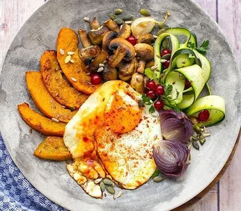 A colorful plate with roasted pumpkin, mushrooms, zucchini ribbons, red onion, sunny side up eggs, and garnished with seeds.