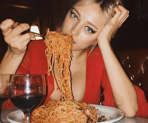 A woman in a red top eating spaghetti with a glass of red wine in front of her at a restaurant.