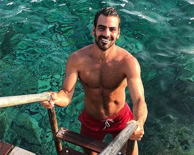 A shirtless man with a beard in red swim trunks climbs a wooden ladder from clear, turquoise water, smiling at the camera.