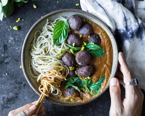 A bowl of noodles with brown sauce, black bean meatballs, fresh basil leaves, topped with crushed nuts on dark surface.