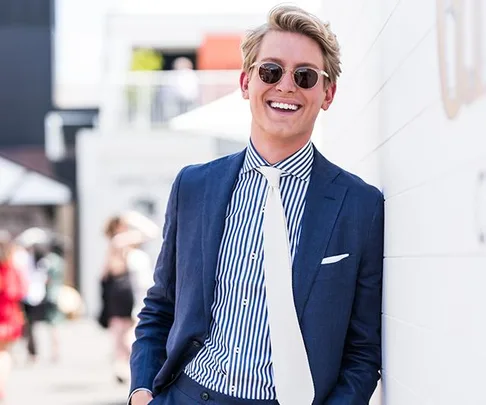 Smiling man in a blue suit, striped shirt, white tie, and sunglasses, leaning against a wall outdoors at Melbourne Cup event.