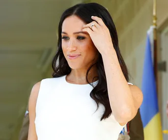 Meghan Markle touching her hair, wearing a white dress and showcasing her engagement ring, in front of a blurred background.