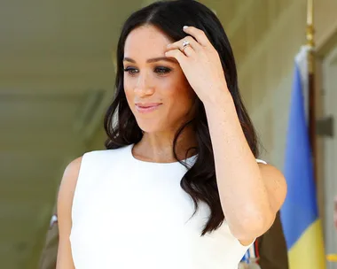 Meghan Markle touching her hair, wearing a white dress and showcasing her engagement ring, in front of a blurred background.