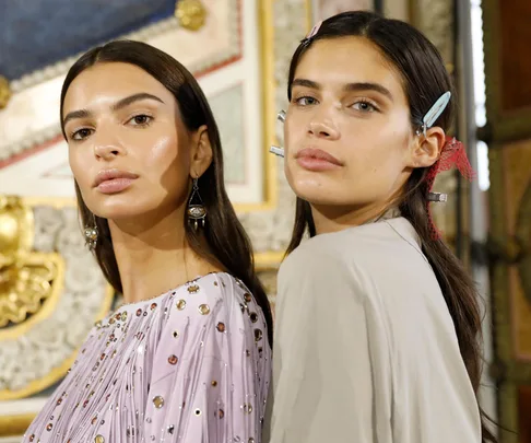 Two women with long hair, wearing elegant outfits, showcasing a no-makeup look at a fashion event.