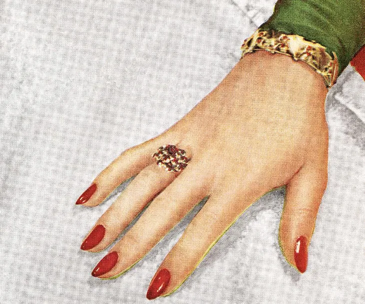 A hand with red nail polish, a jeweled ring, and a gold bracelet on a white background.