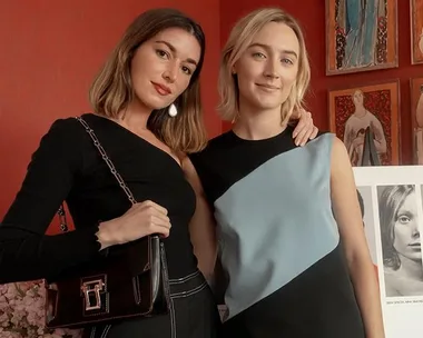 Two women posing together in front of a red wall with art; one wears black, the other a black and blue dress.