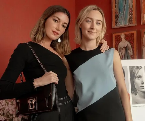 Two women posing together in front of a red wall with art; one wears black, the other a black and blue dress.