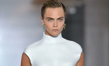 Model with short slicked-back hair in a high-neck white outfit, wearing gold earrings, gazes intently. URL: cara-delevingne-tattoo-18859.