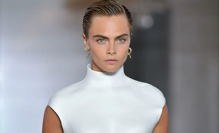 Model with short slicked-back hair in a high-neck white outfit, wearing gold earrings, gazes intently. URL: cara-delevingne-tattoo-18859.