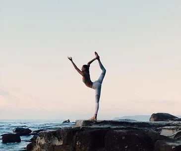 Person performing a yoga pose balancing one leg on rocky shore, ocean and sky in the background.