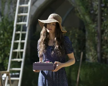 A woman in a blue dress and wide-brimmed hat holding a purple box and standing outdoors in a garden.