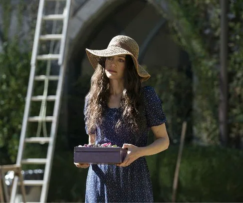 A woman in a blue dress and wide-brimmed hat holding a purple box and standing outdoors in a garden.